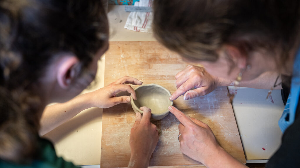 Deux personnes travaillant le modelage d’un petit bol en argile, guidées pas à pas sur une planche de bois.