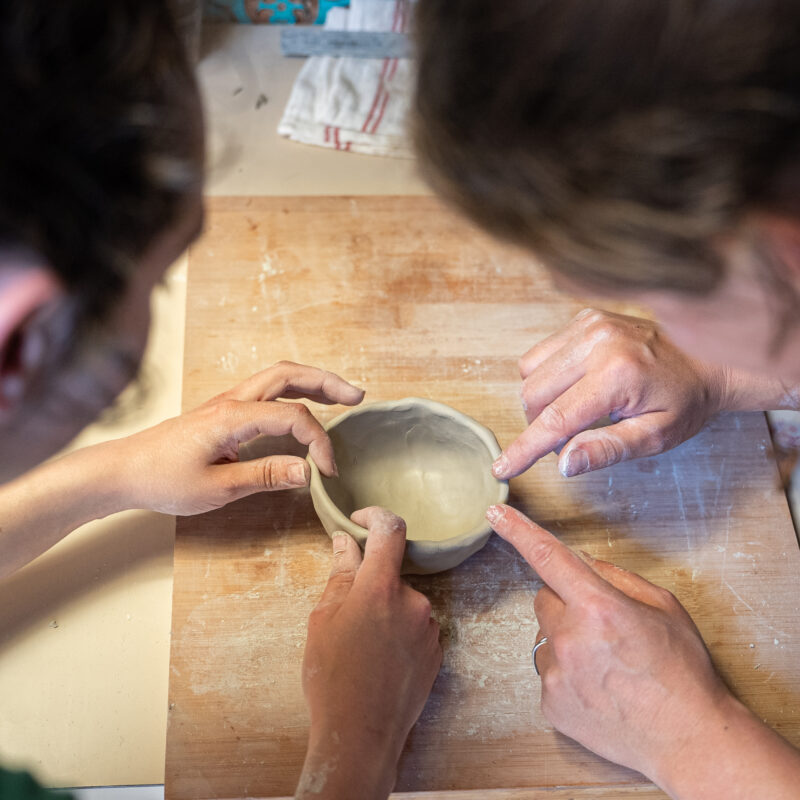 Deux personnes travaillant le modelage d’un petit bol en argile, guidées pas à pas sur une planche de bois.